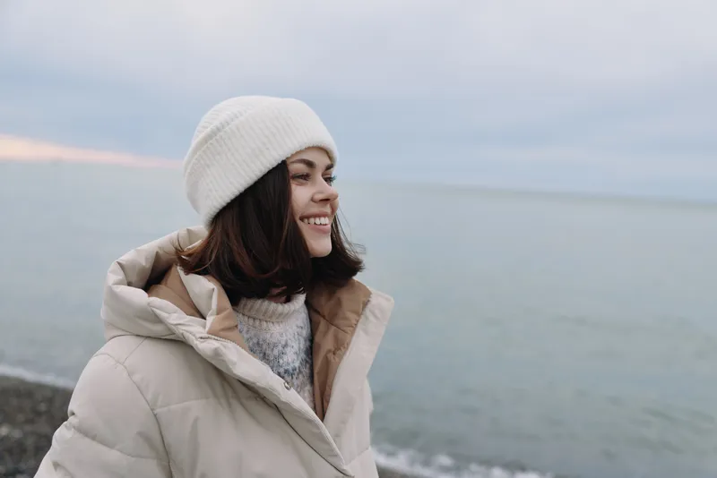 Winter scene featuring a smiling woman in a beige jacket and white beanie by the coast, with sea breeze and soft light creating a calm outdoor mood and casual fashion