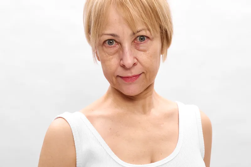 Elderly woman portrait in studio, close view of face and short blonde hair, looking at camera with calm neutral expression, soft light on white background, head and shoulders