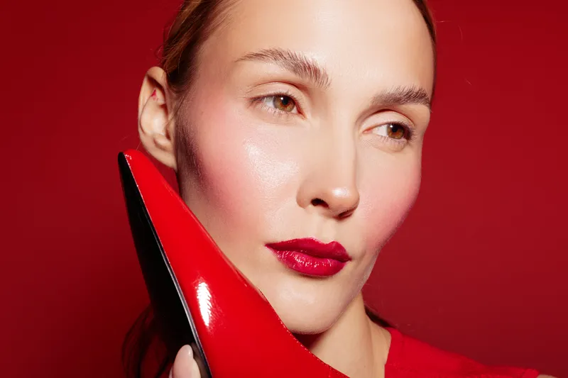 Close-up of a woman with red lipstick holding a red high heel shoe near her face against a red background. Concept of fashion, beauty, style, and elegance with vibrant colors.