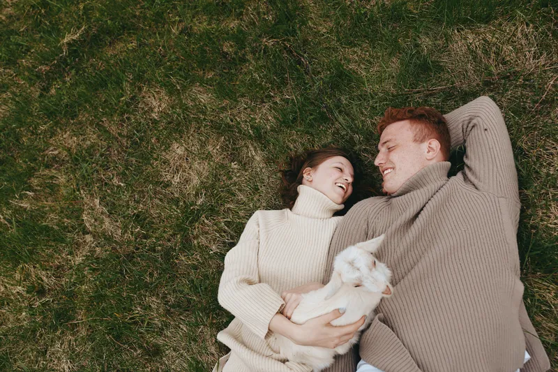 A couple lounges on a grassy lawn, smiling and intertwined, while a white cat rests in their arms. A cozy, intimate moment outdoors, celebrating companionship, happiness, and simple home life in a