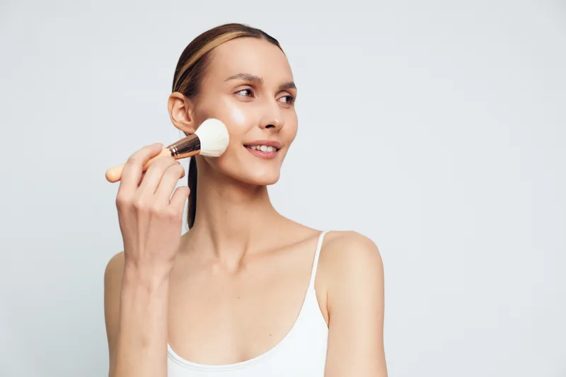 Woman makeup brush cosmetic studio beauty routine grooming, young female applies foundation with careful blending on clear skin, minimal light background, concept for self care
