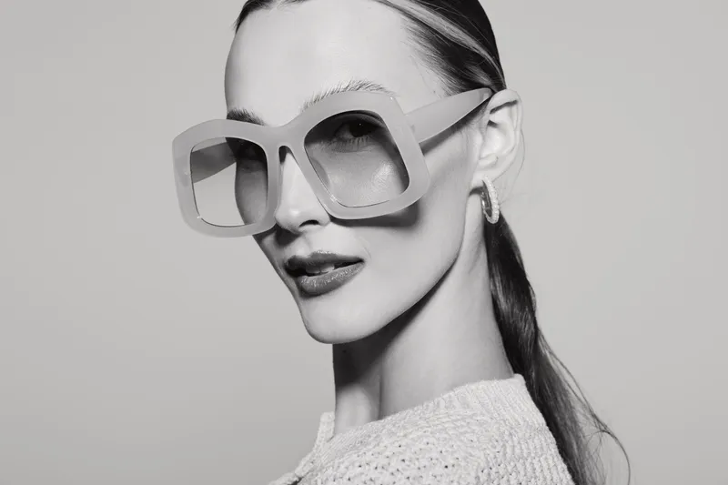Slim white European woman with slicked hair wearing oversized sunglasses and hoop earrings posing with confident expression in studio.