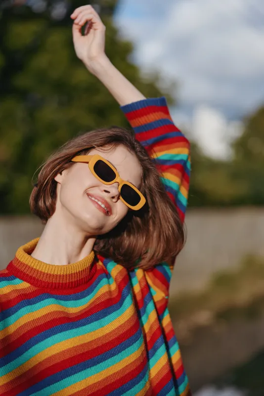woman smile sunglasses rainbow sweater portrait happy fashion outdoors young adult with short hair raising arm enjoying sunny day in colorful knit jumper and joyful relaxed expression