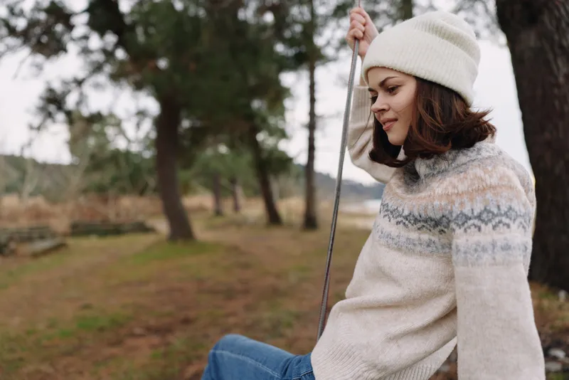 Woman sitting on a swing in a park, wearing a knitted sweater and beanie, relaxing outdoors. Serene casual scene with trees and soft light, perfect for lifestyle, nature, and seasonal moments.