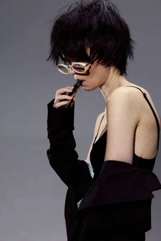 Stylish woman with short hair and sunglasses in black outfit smoking vape indoors