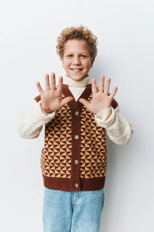 Portrait of a smiling boy standing against a light backdrop, raising both hands in a playful gesture. He wears a geometric brown knitted vest and a cream turtleneck.
