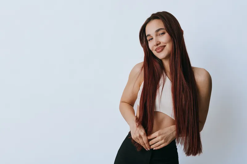 Young woman with long hair wearing sportswear posing with confident smile on white background. Female model standing with hands on waist in casual fitness outfit for lifestyle and health.