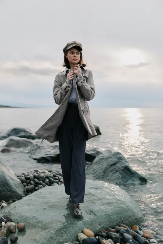 Fashionable woman in a long coat and hat stands on rocky shore near calm sea under cloudy sky. Concept for autumn style, outdoor portrait, and coastal fashion mood.