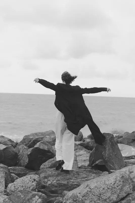 Two figures stand on rugged coastal rocks with arms outstretched toward the open sea, captured in a dramatic black and white scene that conveys freedom, balance, and contemplation by the shore.