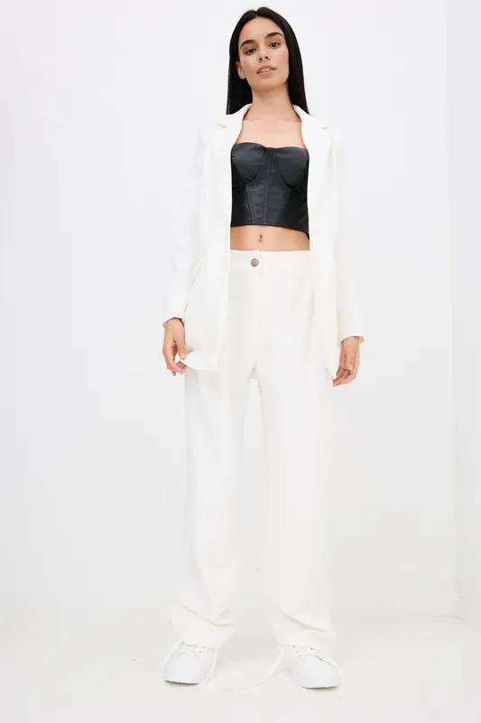Woman wearing white pants and blazer with black cropped top, standing confidently against a plain white background, showcasing modern minimalist fashion style.