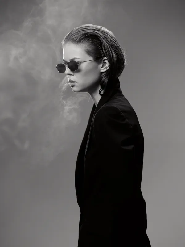 Black and white portrait of a stylish woman in sunglasses with slicked-back hair and smoke in the background.