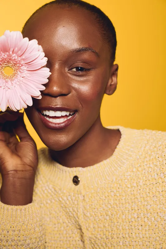 Smiling African woman with dark skin holds pink flower near face, wearing yellow sweater. Close-up portrait on yellow background. Concept of beauty, happiness, and natural skincare.