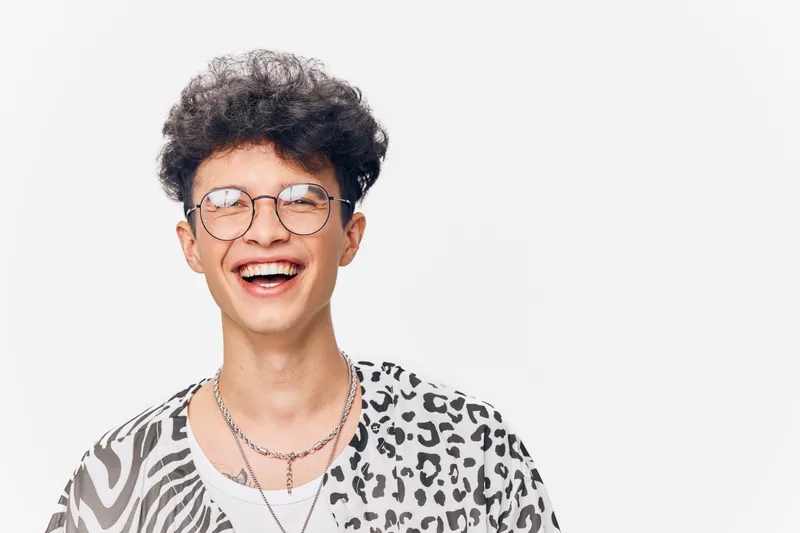 Smiling young man with glasses wearing a patterned shirt, joyful expression, curly hair, white background, casual style, positive mood