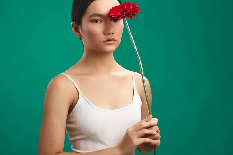 asian woman stands before a plain green background, gently holding a red flower near her face. she gazes softly at the camera, conveying calmness and subtle elegance.