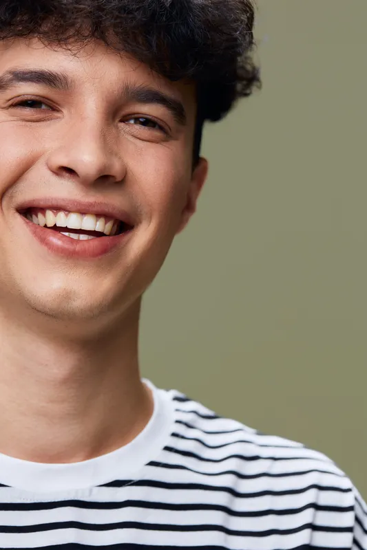 smiling man with curly hair in striped shirt expresses happiness and warmth with natural lighting and blurred green background, casual portrait style