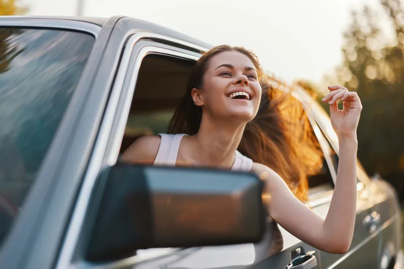 Young woman enjoying a road trip with her head out of the car window, smiling brightly amidst a sunset backdrop, representing freedom and joy of travel.