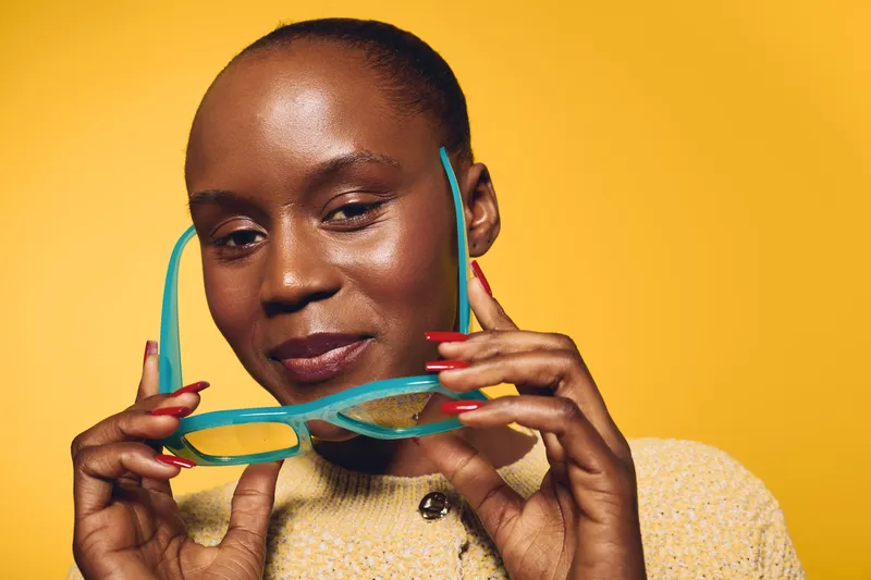 Confident dark skinned African woman with short hair holding turquoise eyeglasses, wearing beige sweater. Yellow background. Concept of beauty, fashion, and self-expression in studio portrait.