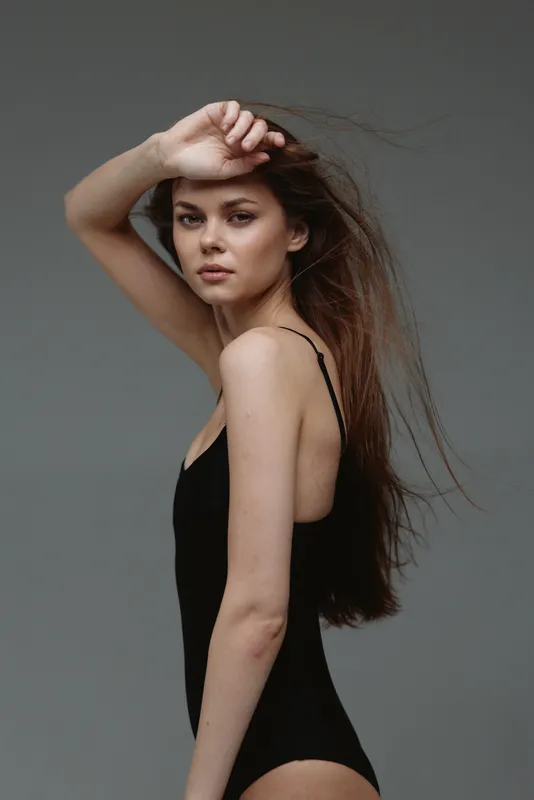 Young woman in black bodysuit posing with hand on forehead against gray background with serious expression.