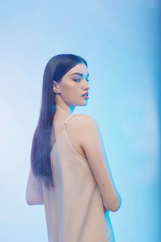 Young woman with long straight hair poses in a beige sleeveless dress against a blue background. Soft lighting highlights her natural beauty and fashion style. Concept of woman beauty fashion.
