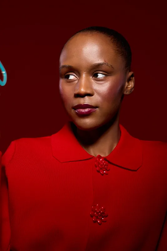 Portrait of a young black woman with dark skin wearing a red sweater against a red background. She looks to the side with a calm expression, highlighting beauty and fashion concepts.