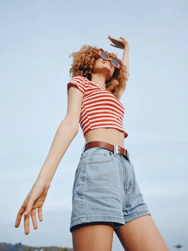 A carefree young woman stands against a clear blue sky, wearing a red striped crop top and denim shorts, arms raised, embodying a bright carefree summer moment.