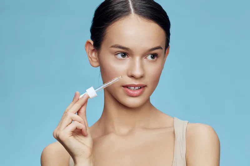 Young woman applying serum on her face with a dropper, showcasing healthy skin and natural beauty against a soft blue background
