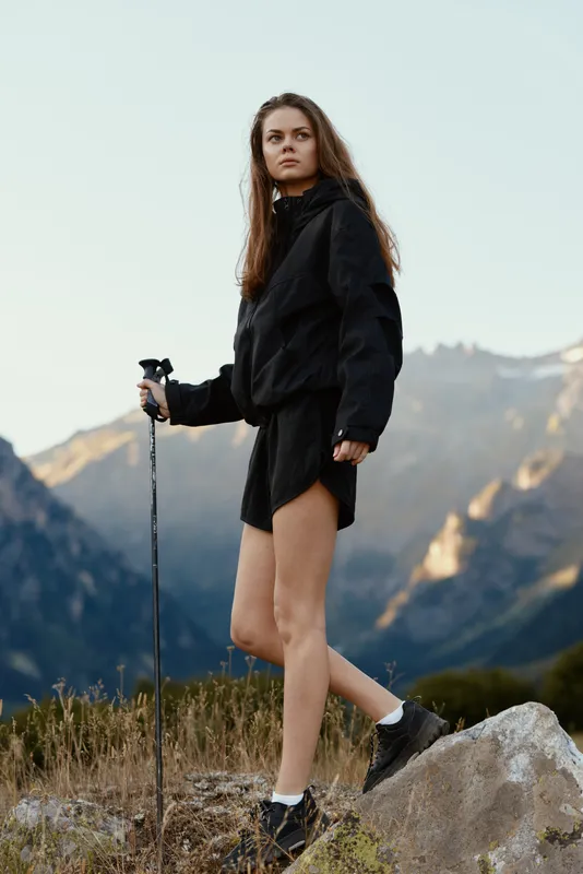 A rugged outdoor enthusiast is seen trekking in a mountain landscape. She wears a dark jacket and shorts, grips a trekking pole, and gazes ahead with focused determination.