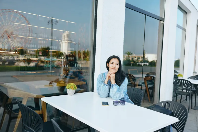 Woman sits at a white outdoor cafe table, thoughtful expression, sunglasses on the table, small plant, glass reflection of a ferris wheel, relaxed urban moment.
