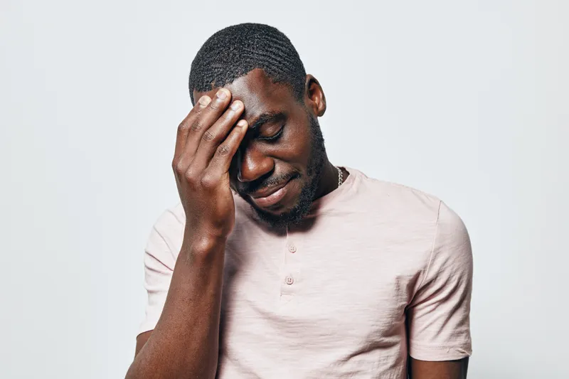 African man with dark skin and short hair touching forehead with closed eyes in stress or headache isolated on white background. Male portrait with emotional expression and casual shirt.