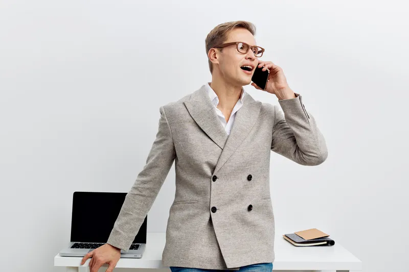 Smiling man talking on mobile phone in office workspace, wearing stylish blazer and glasses, standing next to desk with laptop and notepad, modern business environment, professional lifestyle concept.