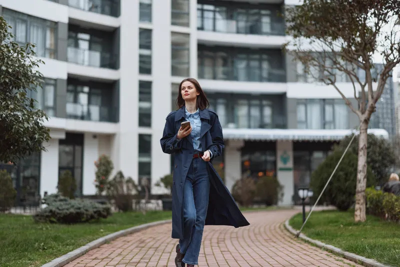 Young woman in a denim jacket and jeans walks along a brick pathway outside a modern building, holding a smartphone and looking ahead with calm confidence.