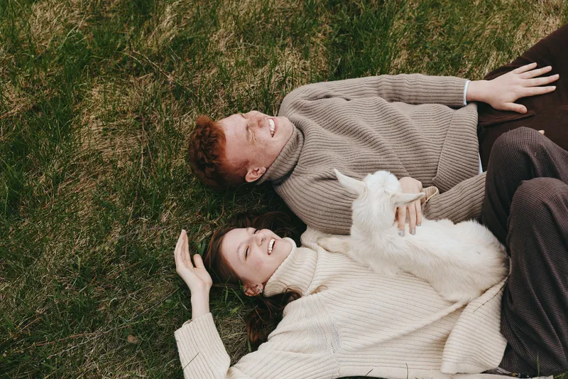 Young couple lying on grass with a white baby goat in a cozy autumn setting wearing knitted sweaters and enjoying a relaxed lifestyle moment outdoors.