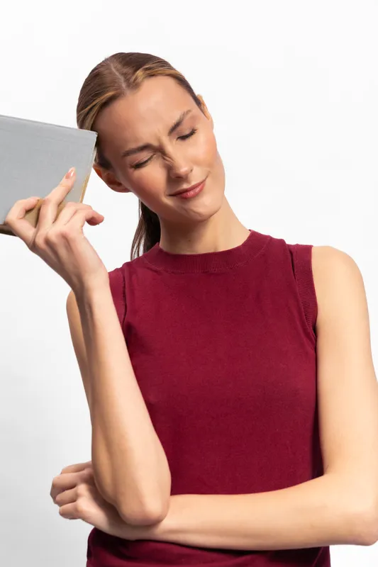 Young woman in red sleeveless top holds a gray book with eyes closed and a frustrated expression. Concept for stress, frustration, and emotional tension in everyday life and study.