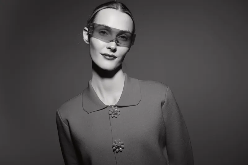 Portrait of a slim Caucasian woman with pale skin wearing sunglasses and a textured jacket in monochrome studio lighting.