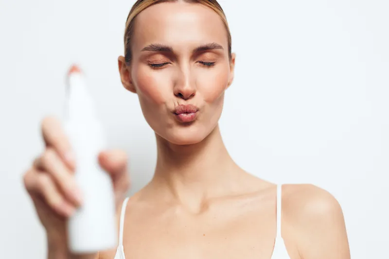 Woman in spa setting close up on face applying skincare serum, holding small bottle, calm expression, soft white background, natural light, beauty treatment today