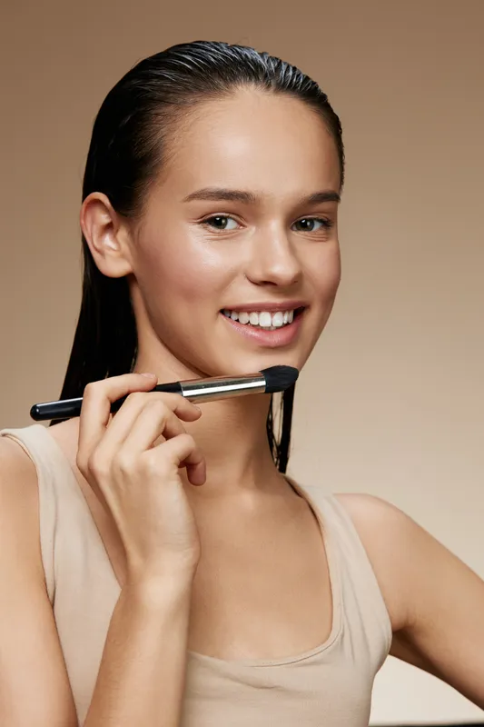 Young female model with a natural look applying makeup with a brush, showcasing a healthy beauty concept, warm background tones emphasizing natural beauty
