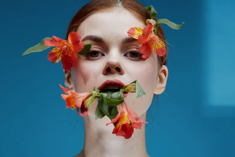 Portrait of a young woman with vibrant orange flowers adorning her face, conveying a sense of beauty and nature against a bright blue background