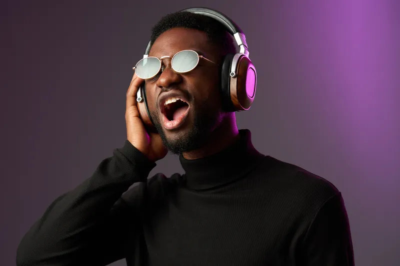 Young Black man wearing sunglasses and headphones, passionately singing against a colorful gradient background, showcasing emotions of joy and music