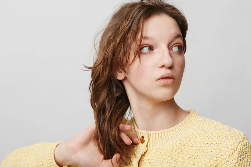 Young woman with wet brown hair wearing a yellow knitted sweater looks to the side on a light grey background. Concept of natural beauty, casual style, and fresh appearance.