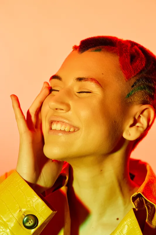 Young woman smiling joyfully with colorful short hair and vibrant yellow jacket on warm orange background, expressing happiness and carefree spirit.