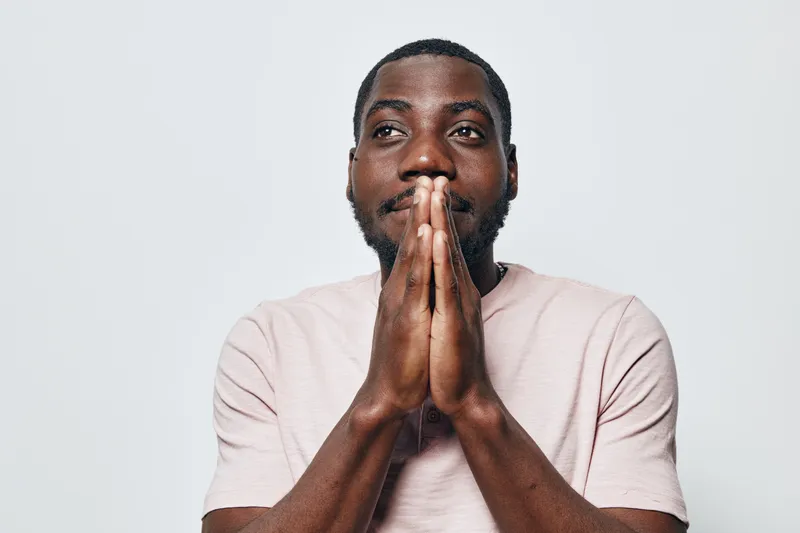 African American man with dark skin and beard wearing light pink shirt, hands pressed together near face, thoughtful expression on white background.