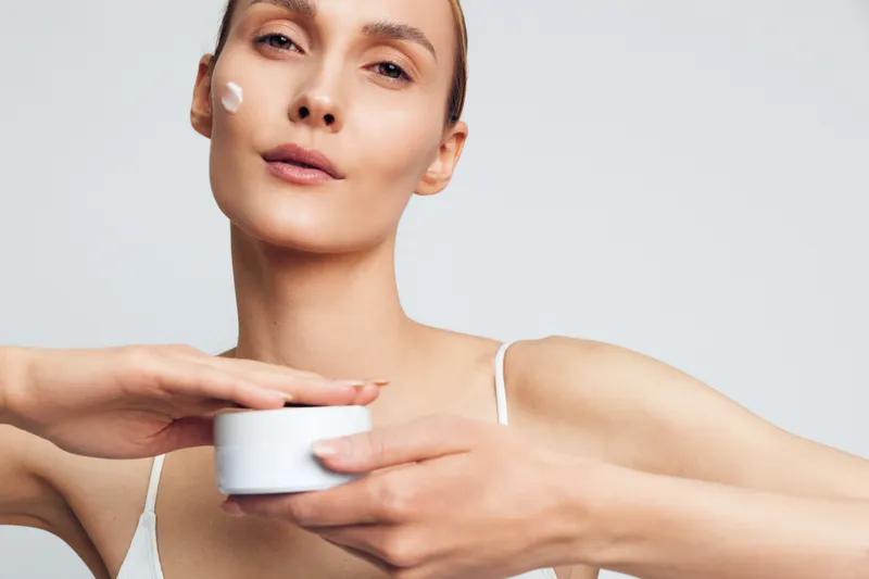 Young woman with clear skin applying white face cream on cheek. Close-up portrait on plain background. Concept of skincare, beauty routine, self-care, and hydration. Soft natural lighting.