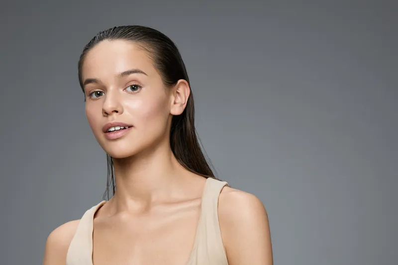 Natural beauty portrait of a young woman with flawless skin and long wet hair, showcasing a minimalist style and serene expression against a grey background