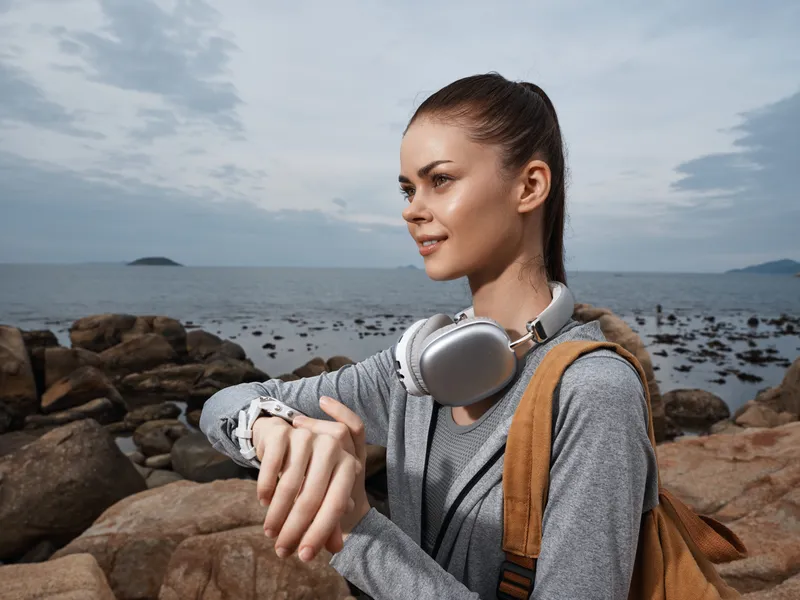 Young woman enjoying nature by the sea, wearing casual sportswear with headphones and a smartwatch, looking at her watch with a thoughtful expression