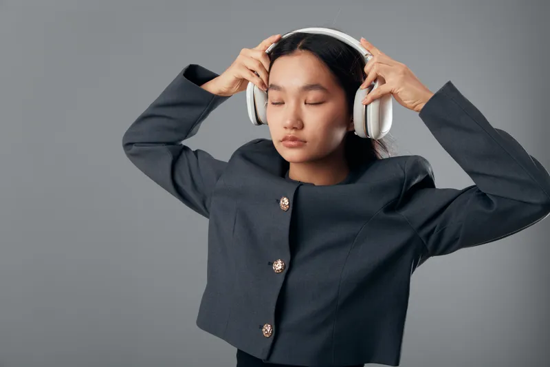 Young woman wearing headphones and closed eyes listening to music with relaxed expression on grey background. Fashion jacket and calm pose for lifestyle and audio experience.
