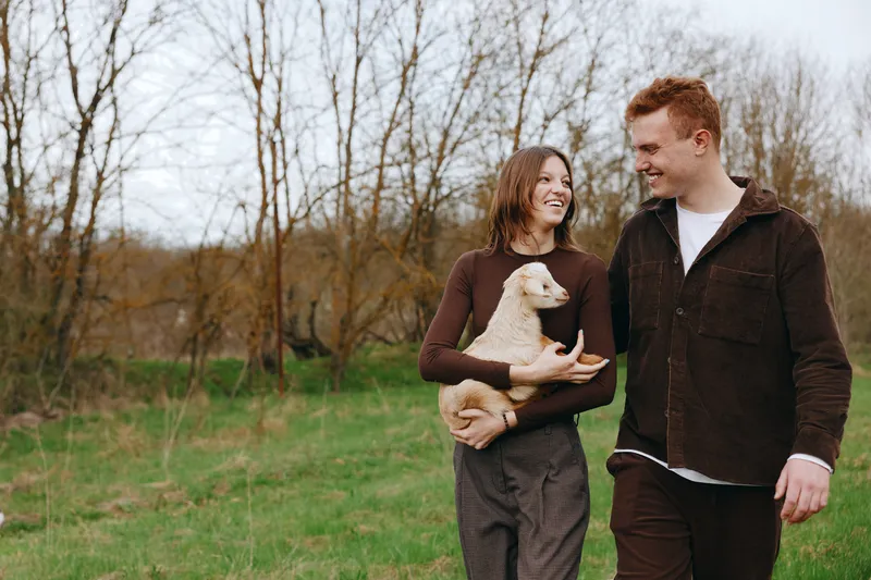 Happy couple outdoors with a small white dog, smiling at each other, embracing in a green field, casual denim and sweaters, candid lifestyle moment. Natural light enhances smiles and warmth.