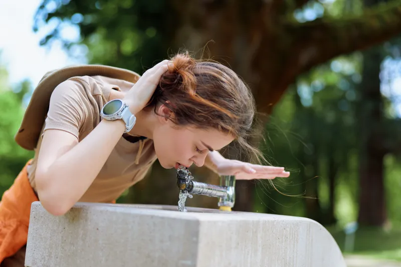 Woman drinking at a fountain in a park with a backpack, leaning over a concrete basin to sip cold water, outdoor summer scene with trees and sunlight, casual youthful refreshment.