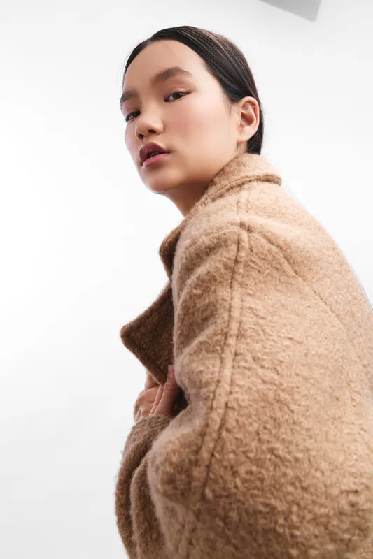 Asian woman wearing a warm tan coat poses in a bright studio, offering a calm, stylish, contemporary portrait that emphasizes confidence and poise.