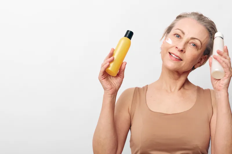 Smiling mature woman with beauty cream on cheeks holds two bottles of skincare products on white background. Concept of facial care, cosmetics, wellness, and self-care for aging skin.