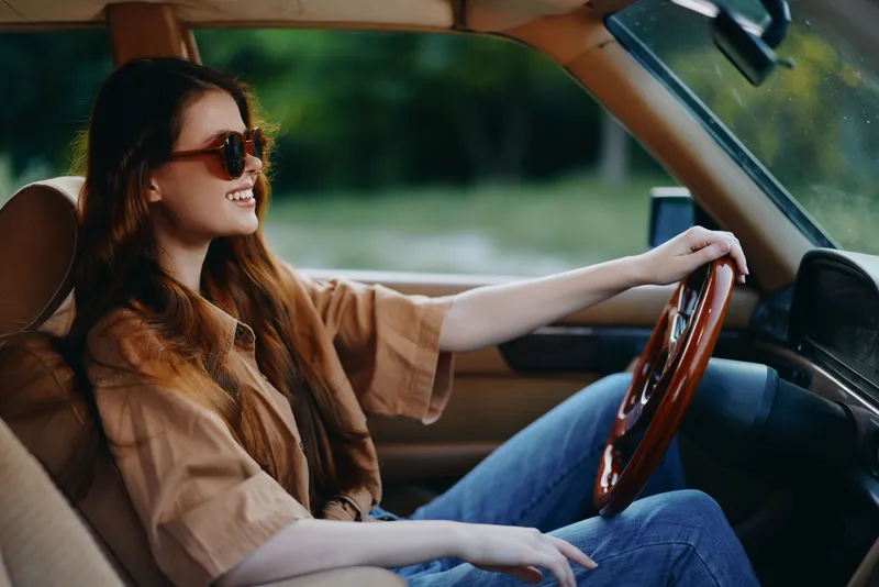 Young woman driving a vintage car, wearing sunglasses and a relaxed outfit, enjoying the summer vibes in a natural green setting.